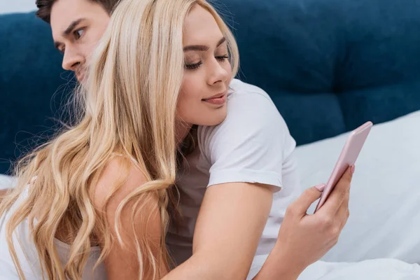 Sorridente giovane donna utilizzando smartphone e abbracciare con il marito a letto, concetto di problema di relazione — Foto stock