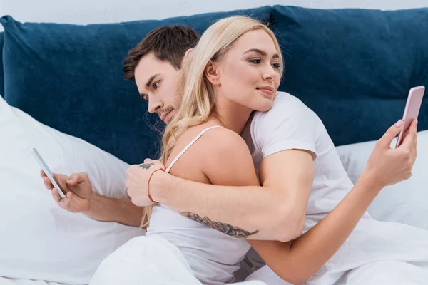 Young couple hugging and using smartphones in bed, relationship problem concept — Stock Photo