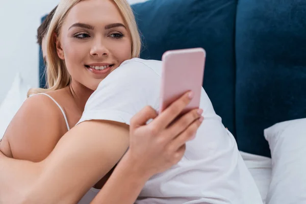 Mujer joven sonriente abrazándose con el novio y usando el teléfono inteligente en la cama - foto de stock