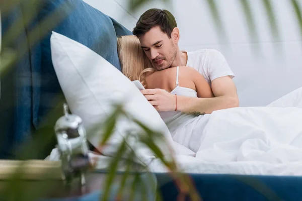 Selective focus of smiling young man using smartphone while lying with girlfriend in bed — Stock Photo