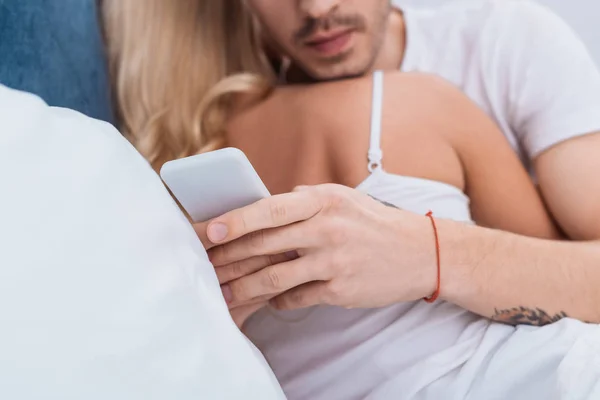 Tiro recortado de homem usando smartphone enquanto abraçando namorada na cama — Fotografia de Stock