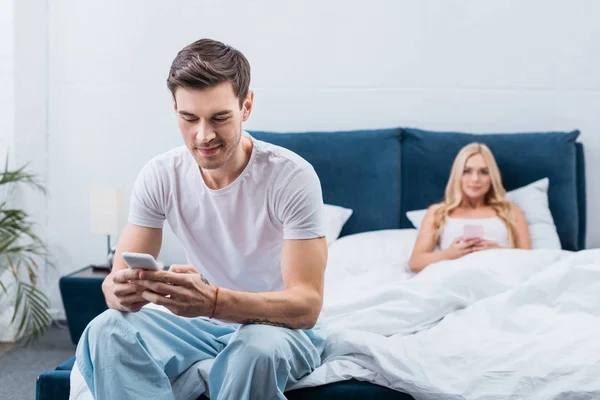 Young woman lying in bed with smartphone and husband using smartphone on foreground in bedroom — Stock Photo