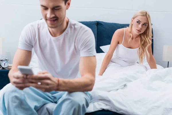 Ревнивая молодая женщина смотрит на парня с помощью смартфона на переднем плане в спальне — стоковое фото