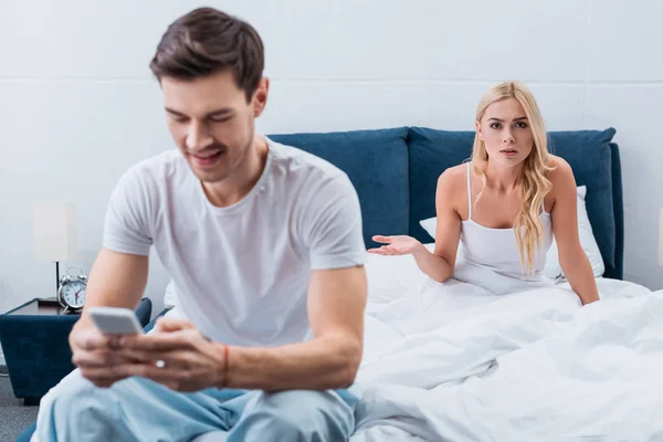 Souriant jeune homme en pyjama en utilisant smartphone et petite amie en colère regardant caméra dans la chambre, concept de méfiance — Photo de stock