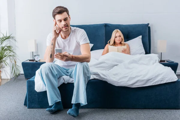 Bonito jovem sentado na cama e usando smartphone enquanto namorada ler livro na cama — Fotografia de Stock