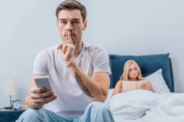 Joven con un gesto de teléfono inteligente para el silencio y mirando a la cámara mientras su novia lee el libro en la cama detrás - foto de stock