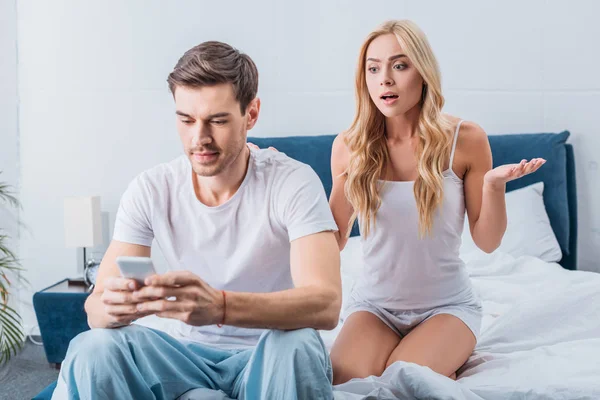 Beautiful shocked woman looking at husband sitting on bed and using smartphone, mistrust concept — Stock Photo