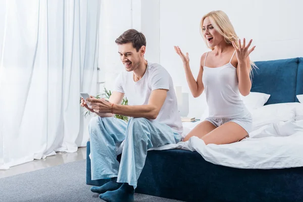 Mujer emocional gritando y sonriendo novio usando smartphone en la cama, concepto de desconfianza - foto de stock