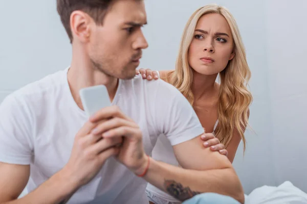 Jeune femme en colère regardant petit ami en utilisant smartphone dans la chambre, concept de problème de relation — Photo de stock