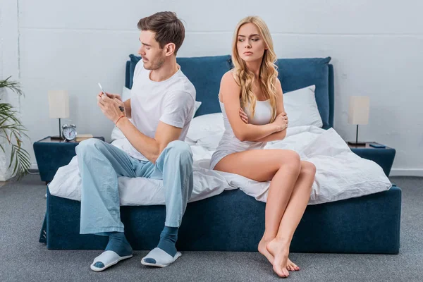 Mujer joven infeliz sentada con los brazos cruzados y mirando al novio usando el teléfono inteligente en la cama - foto de stock