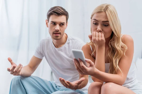 Homme émotionnel parler et regarder jeune femme en utilisant smartphone dans la chambre, concept de méfiance — Photo de stock