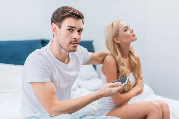 Joven hombre sosteniendo el teléfono inteligente y mirando a la cámara mientras esposa infeliz sentado en la cama, concepto de desconfianza - foto de stock