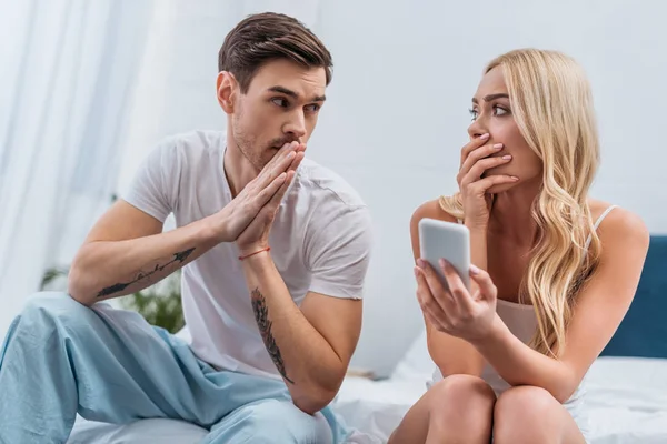 Mujer conmocionada sosteniendo teléfono inteligente y mirando marido confundido sentado en la cama, concepto de desconfianza - foto de stock