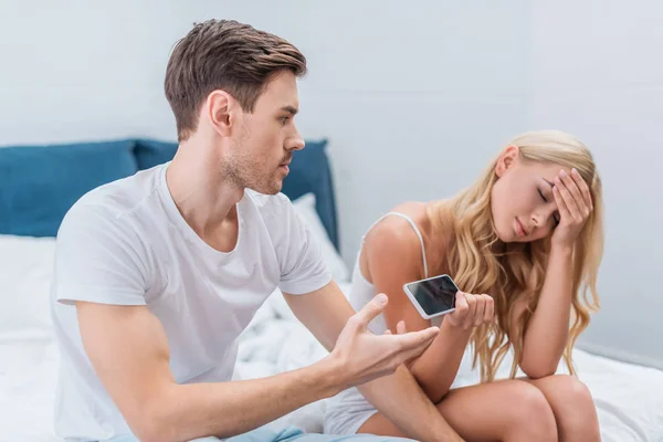 Chateado jovem mulher dando smartphone para namorado sentado na cama, conceito de desconfiança — Fotografia de Stock