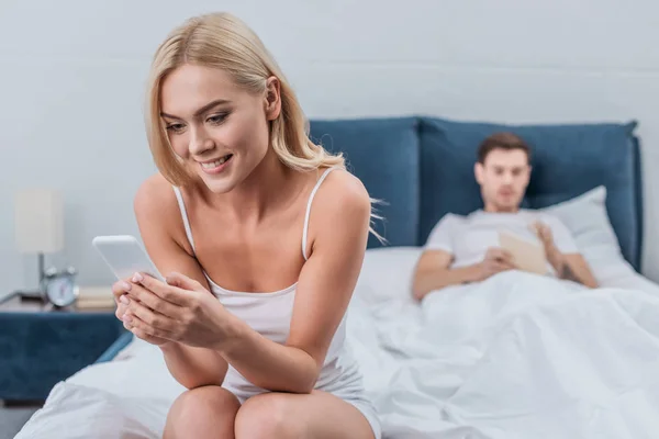 Souriant jeune femme assise sur le lit et en utilisant un smartphone tout petit ami lecture livre derrière — Photo de stock