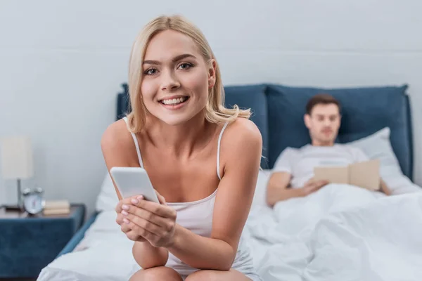 Hermosa mujer joven usando teléfono inteligente y sonriendo a la cámara mientras su novio leyendo libro en la cama detrás - foto de stock