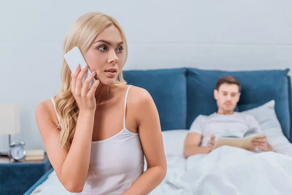 Mujer joven hablando por teléfono inteligente y mirando hacia otro lado mientras su marido lee libro en la cama - foto de stock