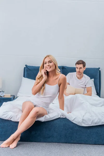 Bella donna sorridente parlando da smartphone mentre geloso fidanzato lettura libro dietro sul letto — Foto stock