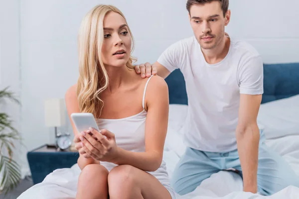 Jealous young man looking at girlfriend using smartphone on bed, mistrust concept — Stock Photo