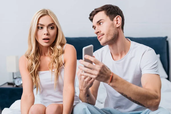 Jealous man showing smartphone to confused girlfriend sitting on bed, distrust concept — Stock Photo