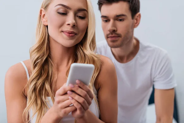 Мужчина смотрит на красивую улыбающуюся жену с помощью смартфона в спальне, концепция недоверия — стоковое фото