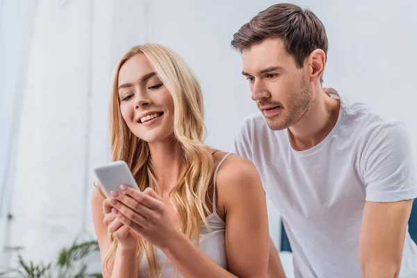 Jealous young man looking at beautiful smiling girlfriend using smartphone in bedroom — Stock Photo