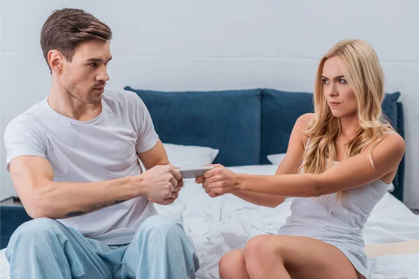 Jovem casal zangado lutando por smartphone na cama, conceito de desconfiança — Fotografia de Stock