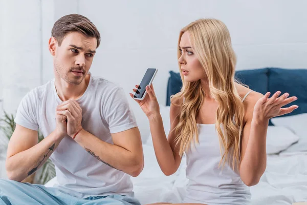 Confused woman showing smartphone to suspicious boyfriend in bedroom — Stock Photo