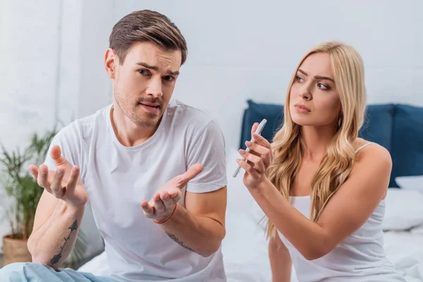Upset woman showing smartphone to emotional suspicious husband sitting on bed — Stock Photo