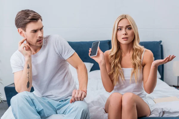 Confused woman holding smartphone and looking at camera while sitting with boyfriend on bed — Stock Photo