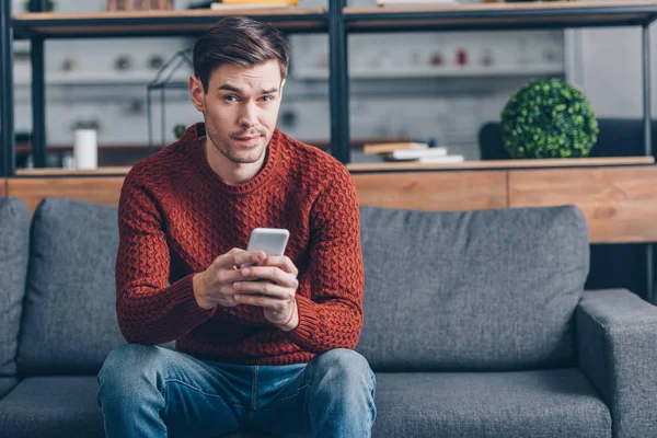Suspicious young man holding smartphone and looking at camera while sitting on couch at home — Stock Photo