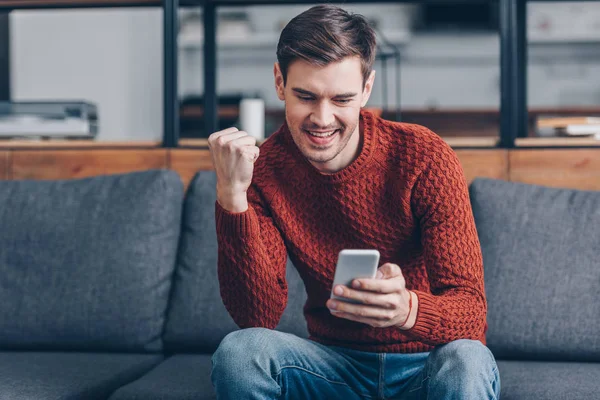 Excited young man sitting on couch and using smartphone at home — Stock Photo