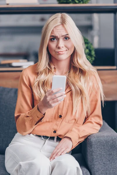 Junge Frau mit Smartphone runzelt die Stirn und blickt in Kamera drinnen — Stockfoto