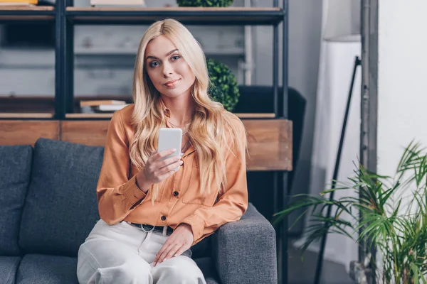 Красива блондинка використовує смартфон і посміхається на камеру, сидячи на дивані вдома — стокове фото