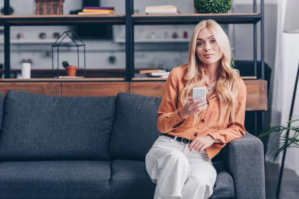 Attractive blonde girl using smartphone and smiling at camera while sitting on couch at home — Stock Photo