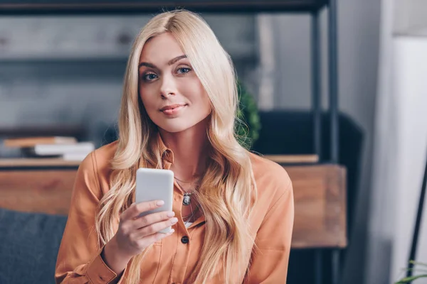 Молодая женщина с помощью смартфона и улыбаясь перед камерой в помещении — стоковое фото