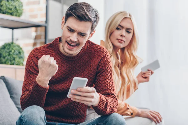 Pareja joven usando teléfonos inteligentes en casa, hombre emocionado sacudiendo puño, concepto de desconfianza - foto de stock