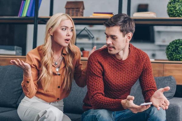 Emocional pareja molesta sentado en el sofá y peleando por el teléfono inteligente, concepto de desconfianza - foto de stock