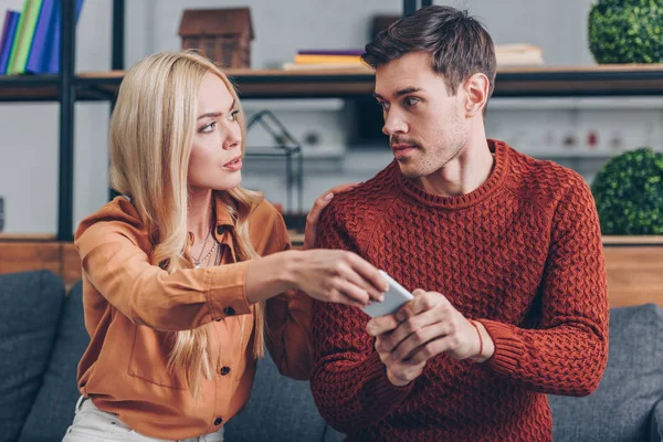 Emocional joven pareja sosteniendo el teléfono inteligente y mirándose el uno al otro, concepto de celos - foto de stock