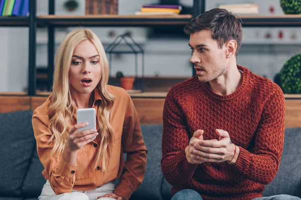 Emotional young man looking at shocked girlfriend using smartphone on couch, jealousy concept — Stock Photo