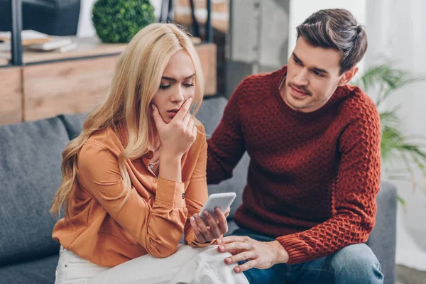 Young man looking at upset girlfriend using smartphone on couch, distrust concept — Stock Photo