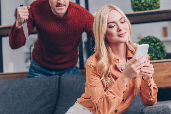 Recortado disparo de hombre agresivo sosteniendo cuchillo mientras sonríe novia usando teléfono inteligente en el sofá, concepto de celos - foto de stock