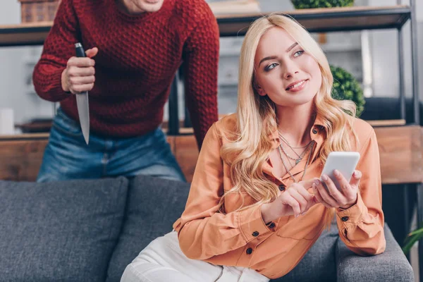 Tiro recortado de homem agressivo segurando faca enquanto sorrindo esposa usando smartphone no sofá, conceito de ciúme — Fotografia de Stock