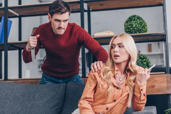Aggressive man with knife looking at girlfriend using smartphone on couch, betrayal concept — Stock Photo