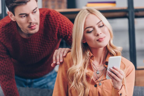 Mari regardant belle femme souriante en utilisant un smartphone à la maison, concept de jalousie — Photo de stock