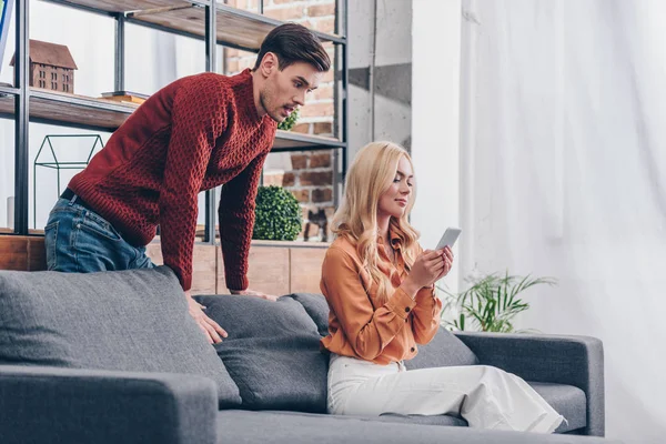Emotional young man looking at smiling girlfriend sitting on couch and using smartphone, jealousy concept — Stock Photo