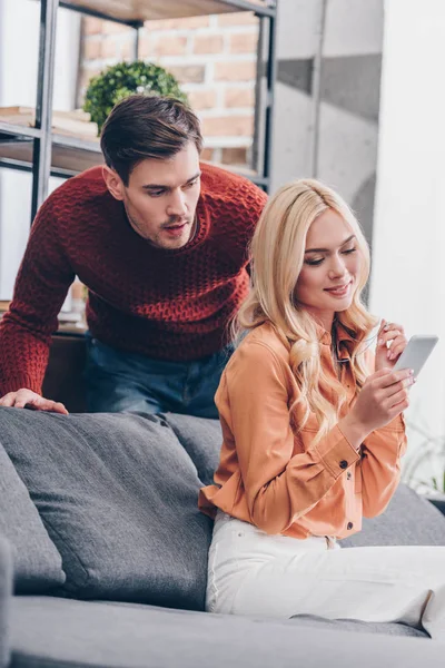 Ревнивый эмоциональный мужчина смотрит на улыбающуюся молодую жену с помощью смартфона дома, концепция недоверия — стоковое фото
