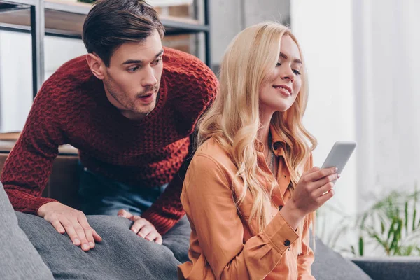 Шокированный ревнивый мужчина смотрит на улыбающуюся молодую женщину с помощью смартфона дома, концепция недоверия — стоковое фото