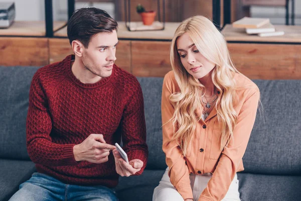 Hombre joven celoso mostrando teléfono inteligente a mujer joven confundida sentada en el sofá - foto de stock