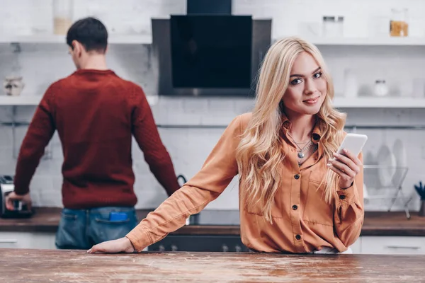 Hermosa mujer joven utilizando el teléfono inteligente y sonriendo a la cámara mientras el marido de pie en la cocina detrás - foto de stock